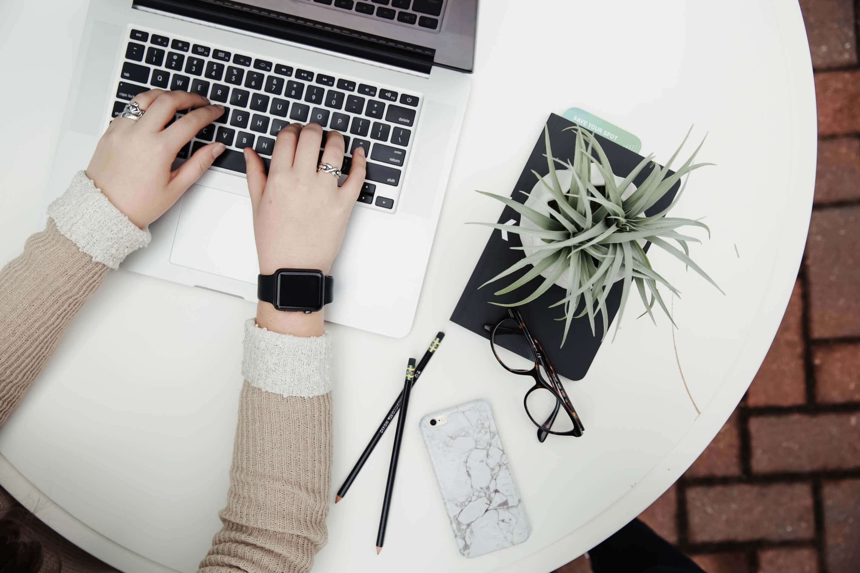 Wieso Managed Services? Hände einer Person auf einem Laptop, welcher auf einem weißen, runden Tisch steht. Neben dem Laptop steht eine Pflanze auf einem Buch.